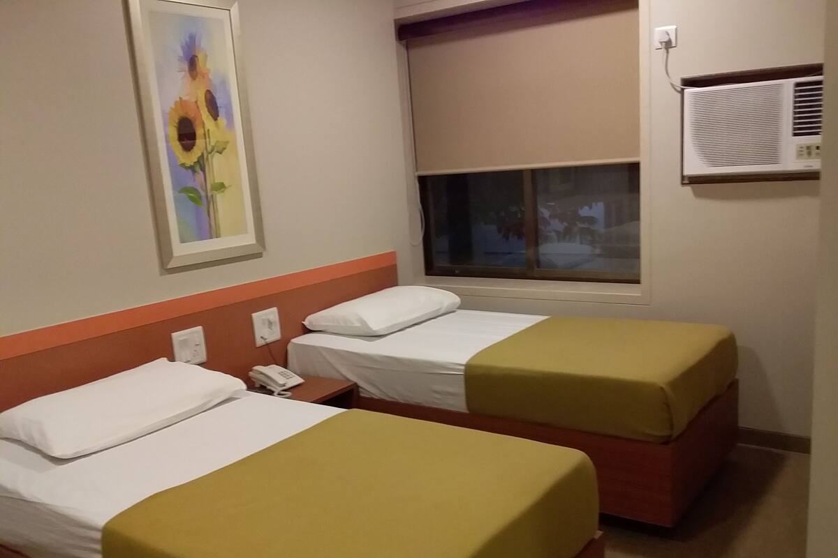 A hotel room with two beds, each with a white mattress and a yellow bedspread. There is a painting of sunflowers on the wall, a window with a view of the outside, and an air conditioning unit mounted on the wall.