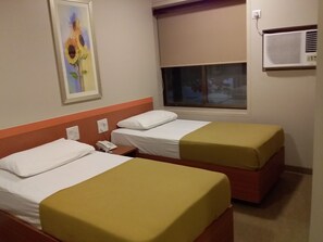 A hotel room with two beds, each with a white mattress and a yellow bedspread. There is a painting of sunflowers on the wall, a window with a view of the outside, and an air conditioning unit mounted on the wall. - Hotel Sai Palace Express (Rahata)