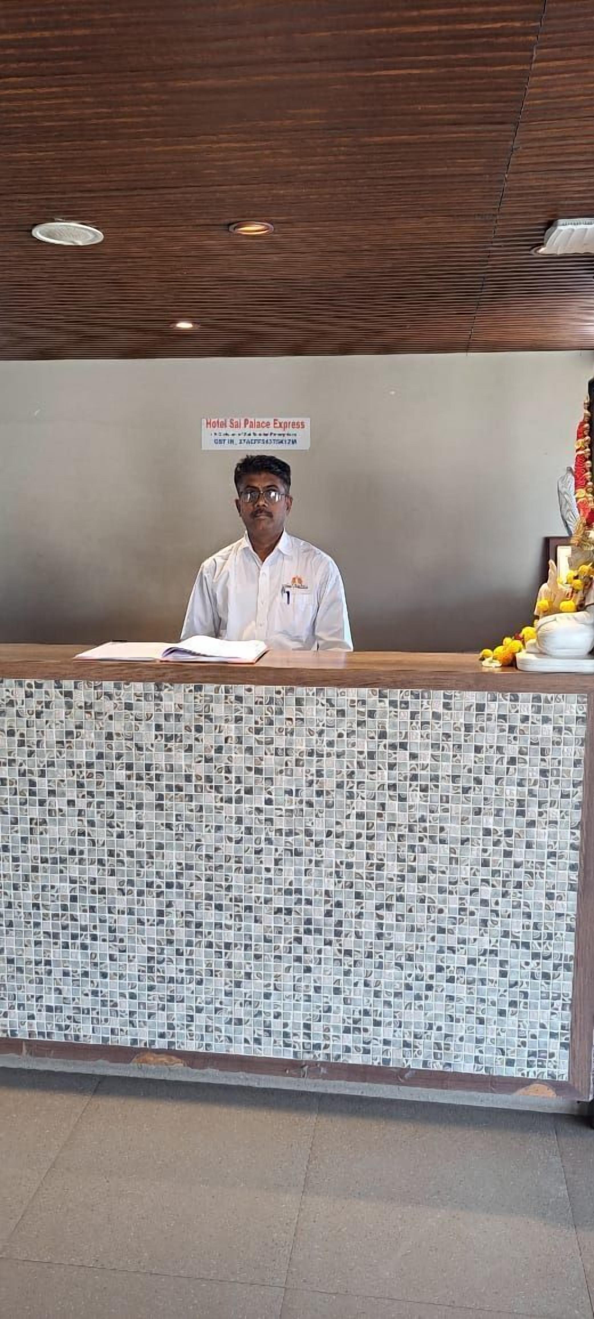 A hotel reception desk with a mosaic tile design, a sign above indicating the hotel name, and a small statue on the right.