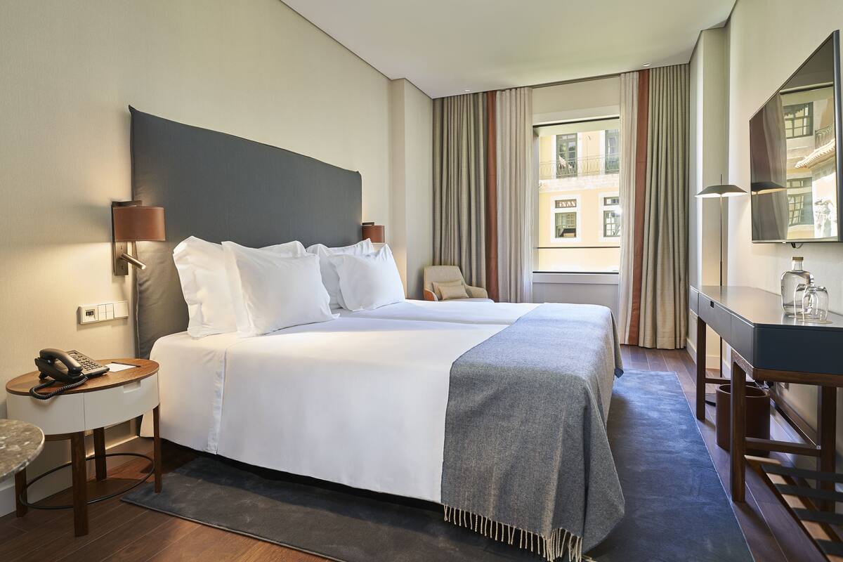 A hotel room with a bed, bedside table, lamp, and a desk with a chair.