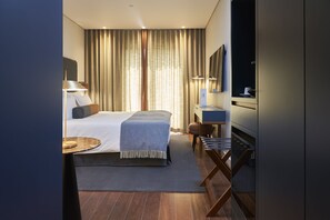 A modern hotel room with a neatly made bed, a desk with a lamp, a chair, and a TV mounted on the wall.
