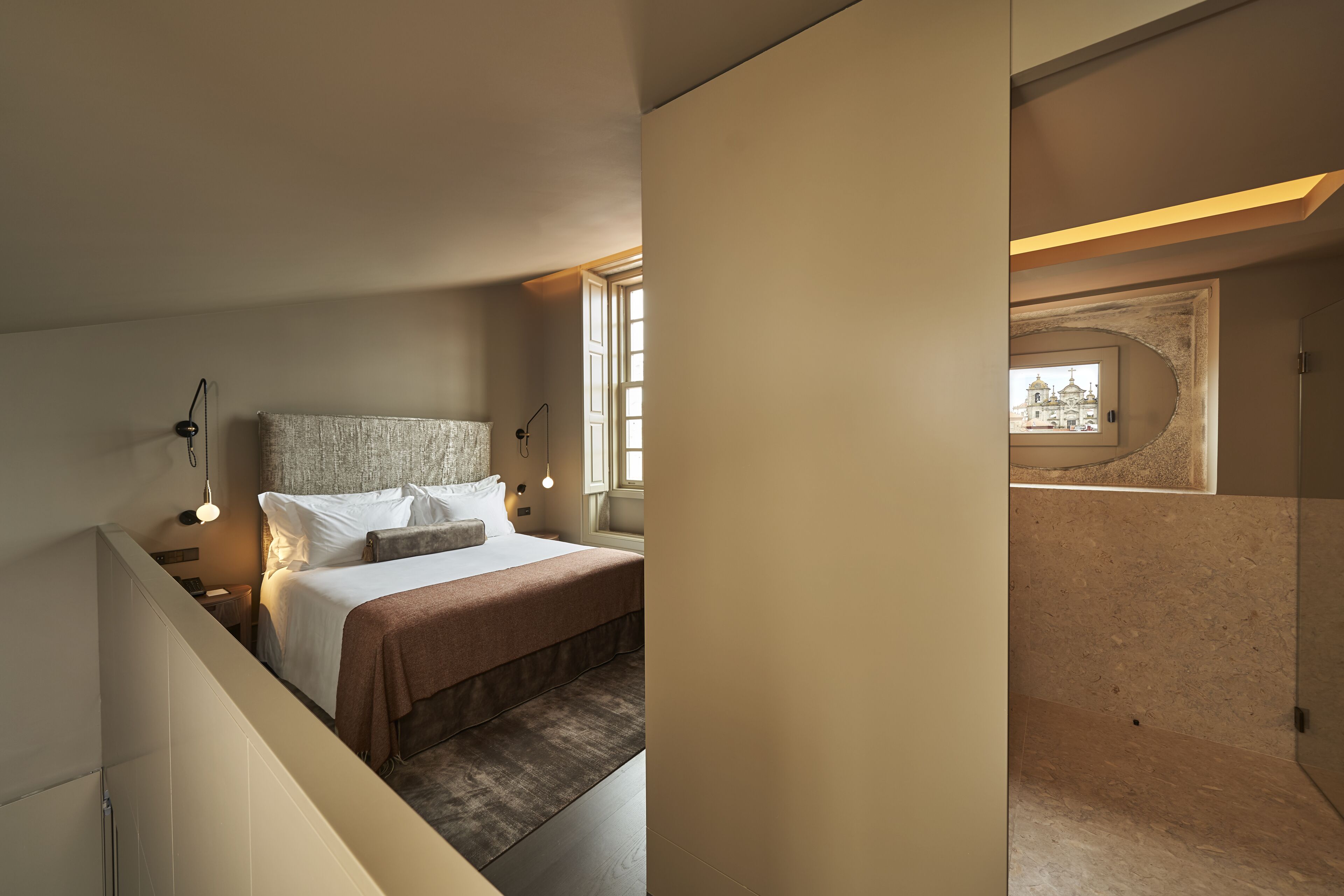 A modern hotel room with a large bed, a view of a building through a window, and a small balcony.