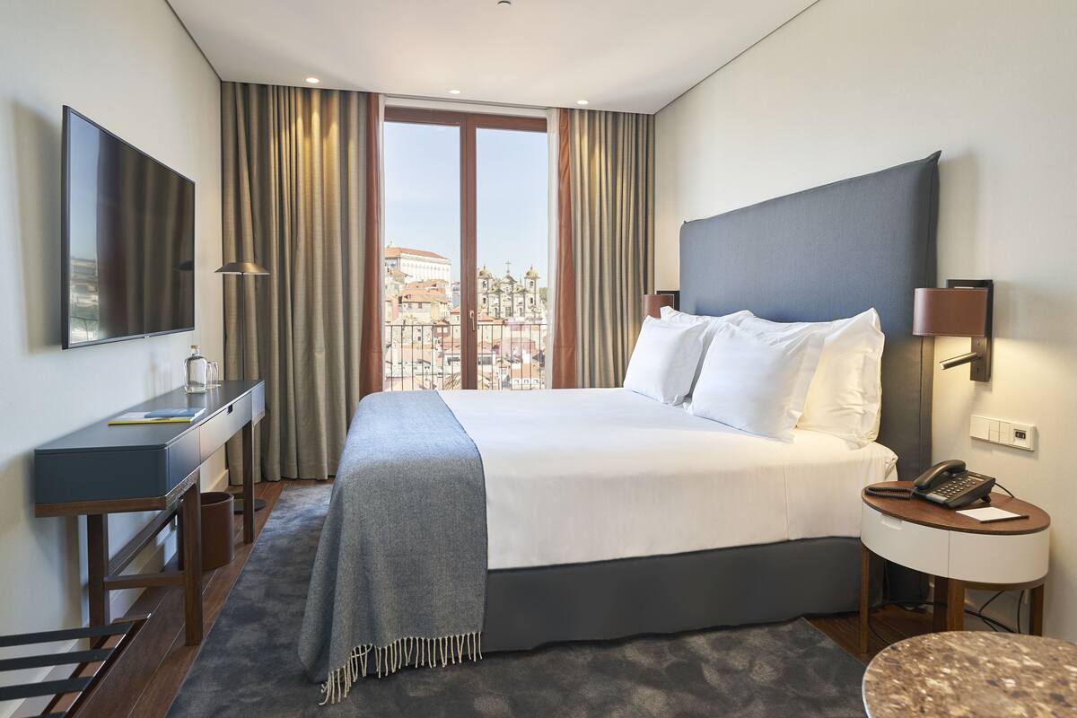 A modern hotel room with a large bed, a desk, a chair, a small table, and a view of the city through the window.