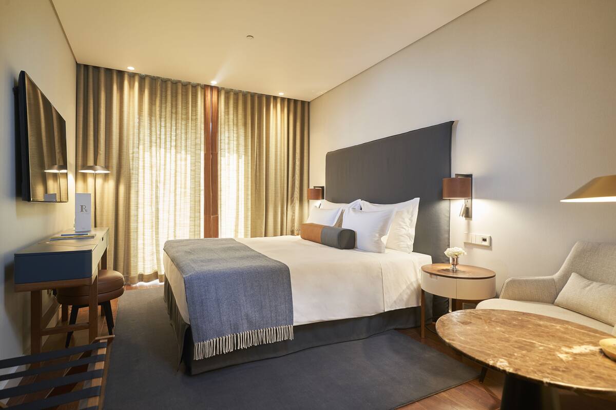 A modern hotel room with a large bed, a desk, a chair, a round coffee table, and a sofa.