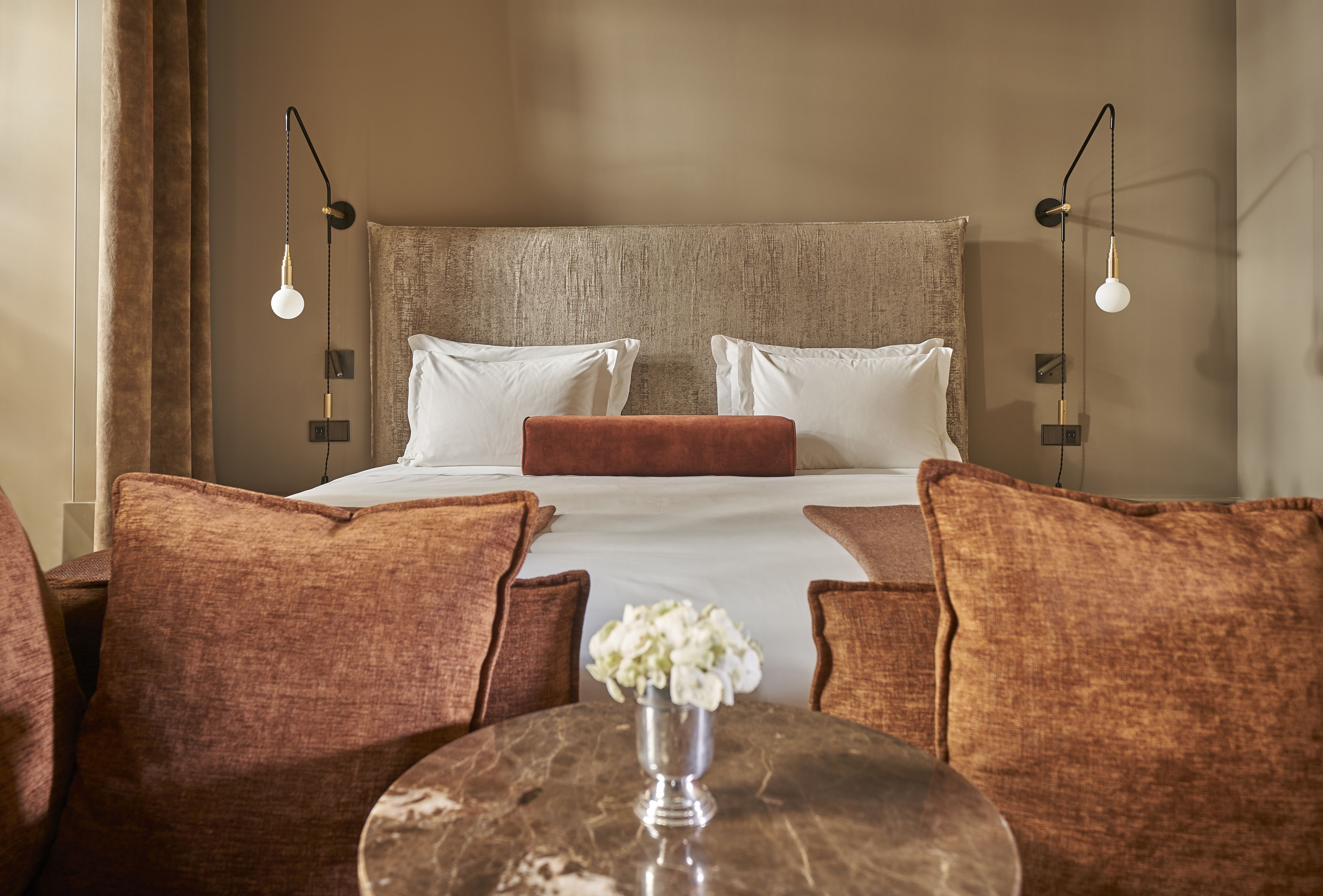 A hotel room with a large bed, two bedside tables, a round table with a vase, and brown upholstered chairs.