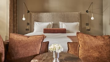 A hotel room with a large bed, two bedside tables, a round table with a vase, and brown upholstered chairs.