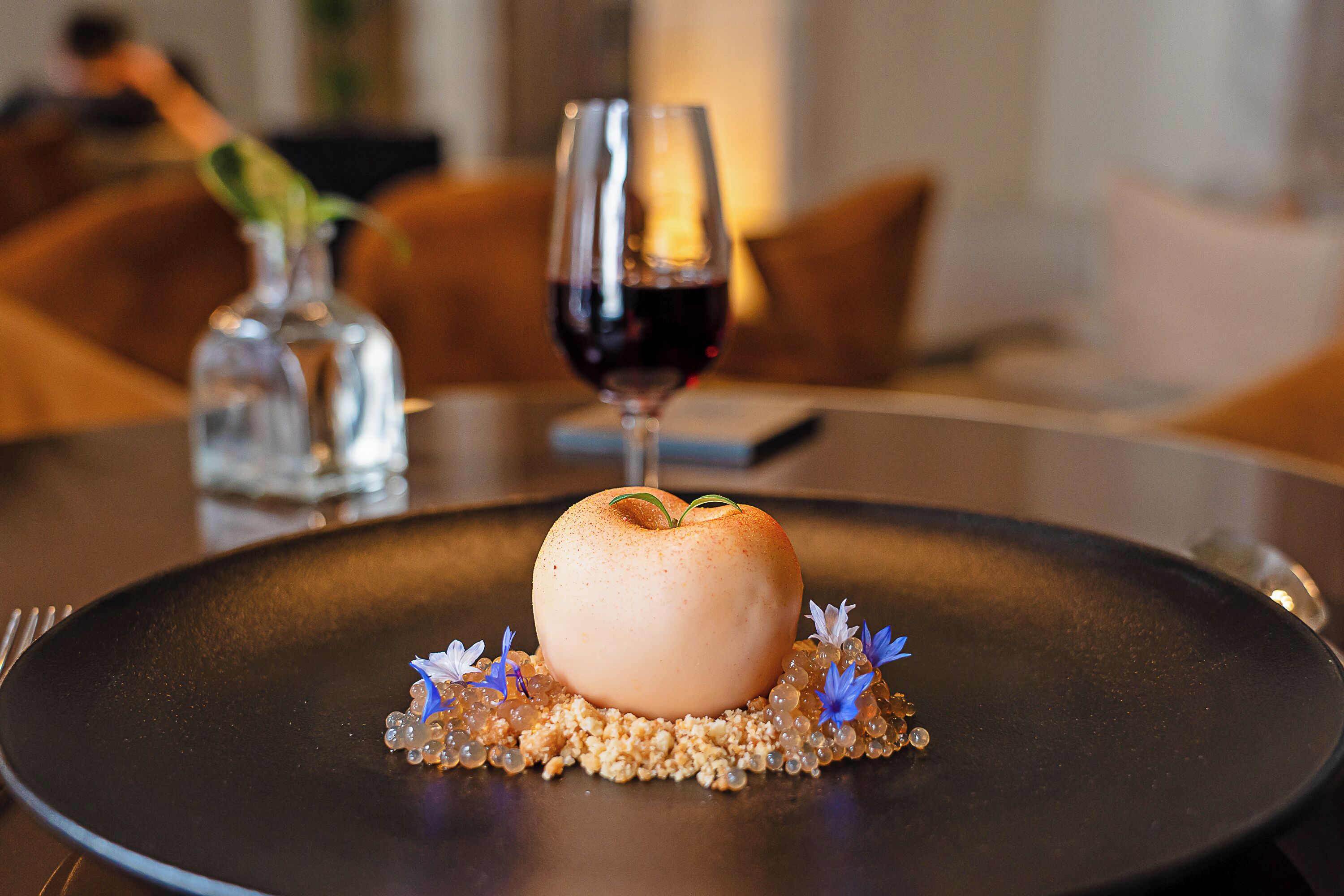 Un postre en un plato con una copa de vino tinto y un jarrón con flores.