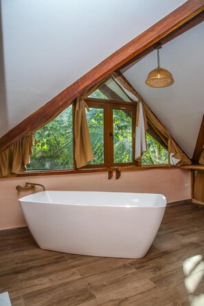 A room with a large bathtub, wooden beams, a window with curtains, and a hanging light fixture. - La Brisa Tranquila - Hostel (Santa Marta)