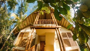Une maison traditionnelle en bois, dotée d’un escalier menant à un balcon, et entourée d’une végétation tropicale luxuriante.