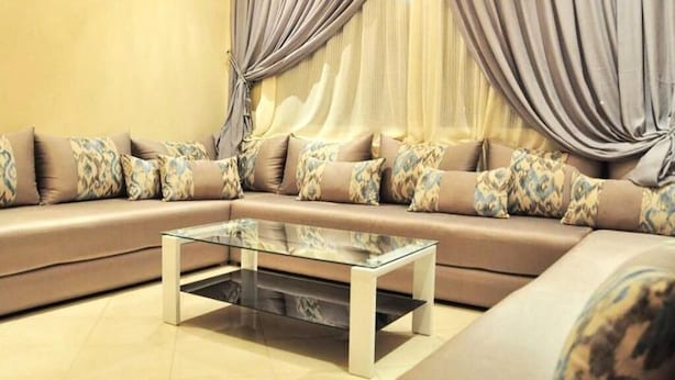 Un salon moderne avec un canapé d’angle beige, des coussins décoratifs à motifs et une table basse en verre.