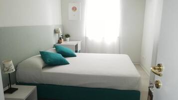 A bedroom with a bed, a nightstand, a lamp, a small table with a plant, a picture on the wall, and a door with a brass handle.