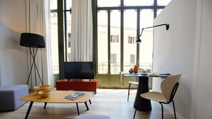A modern living room with a flat-screen TV, a wooden coffee table, a sofa, a dining table with a chair, and a large window with a view of buildings.