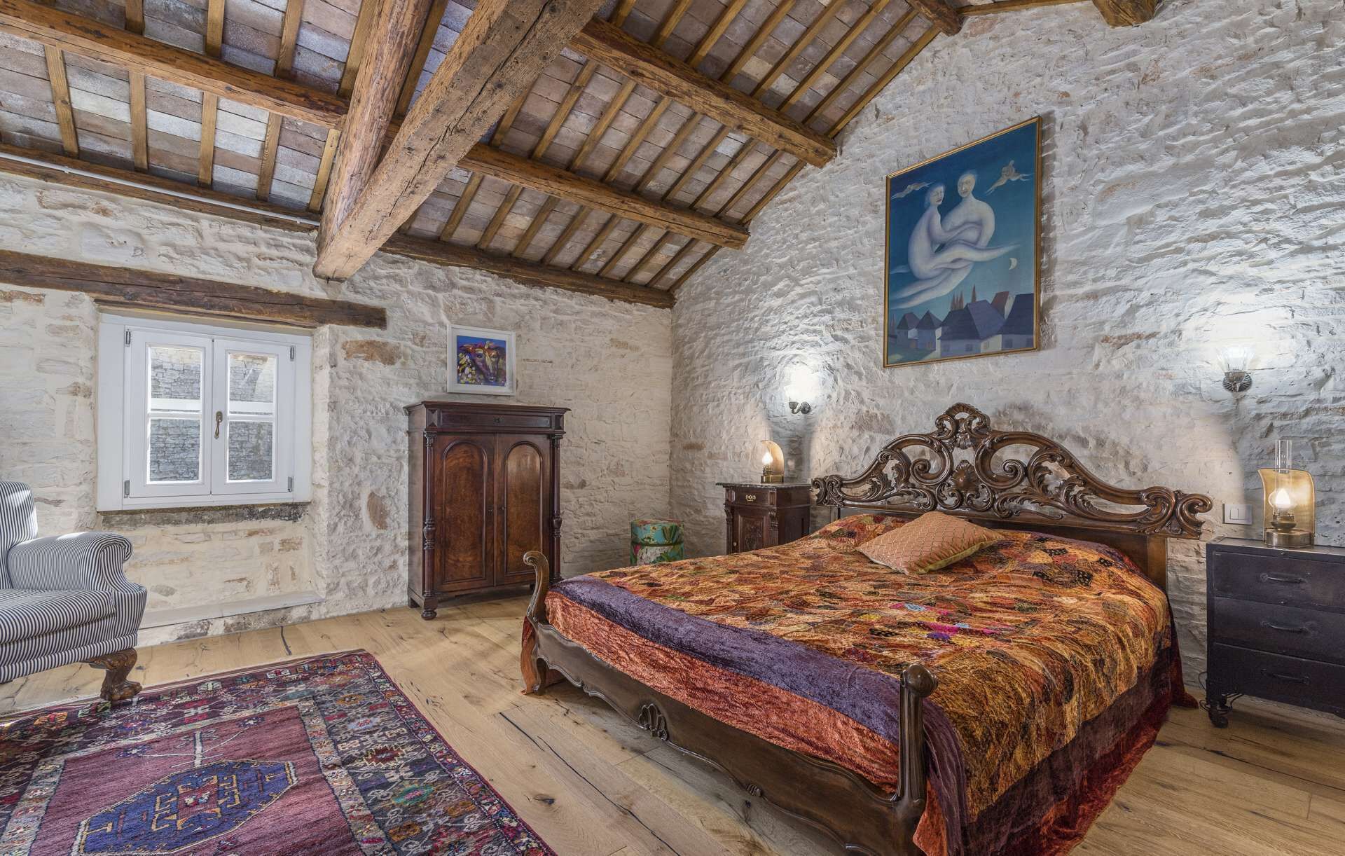 Een rustieke slaapkamer met houten plafond, een groot bed, een stenen muur en een raam met witte luiken.