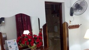 Une chambre d’hôtel avec deux lits, un bureau en bois avec un miroir, un arrangement floral rouge, un ventilateur fixé au mur et une petite table avec une lampe.
