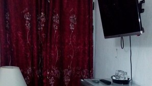 Une petite pièce avec un téléviseur fixé au mur, une petite table avec une lampe, un seau rouge et un sol carrelé.