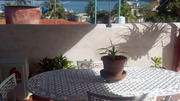 Une terrasse extérieure avec une table ronde en métal et deux chaises, un pot de fleurs, et une vue sur la mer et les montagnes en arrière-plan.
