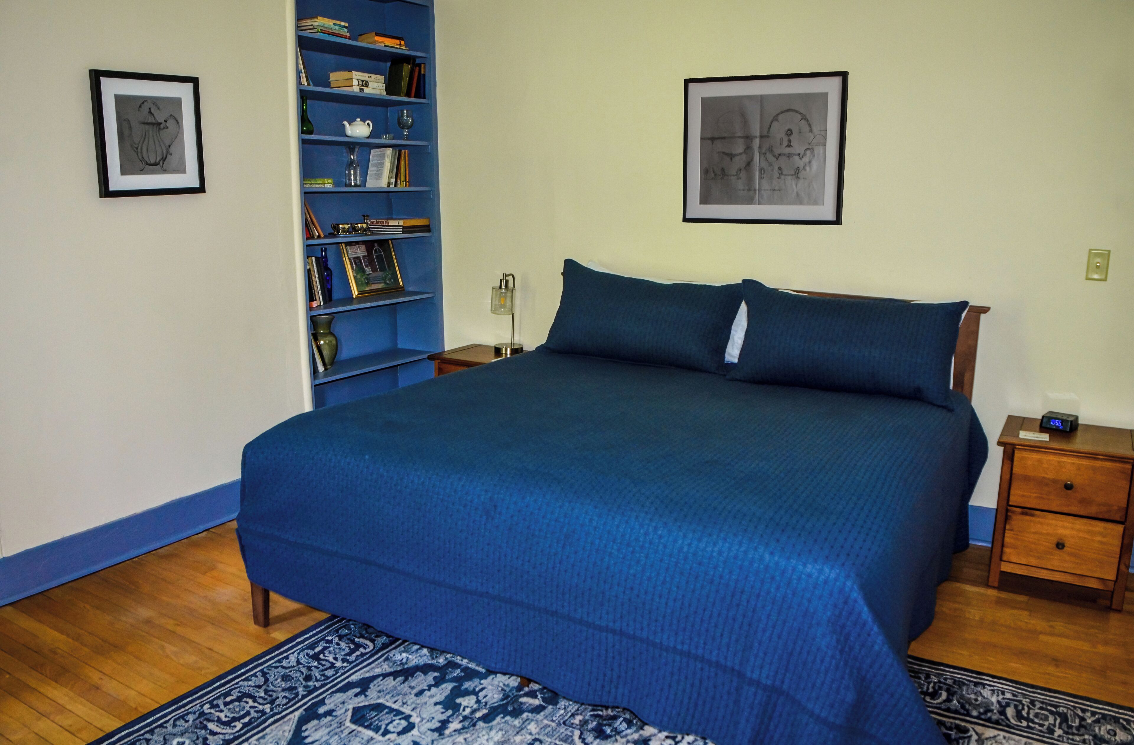 A bedroom with a large bed, a bookshelf, a nightstand, and two framed pictures on the wall.