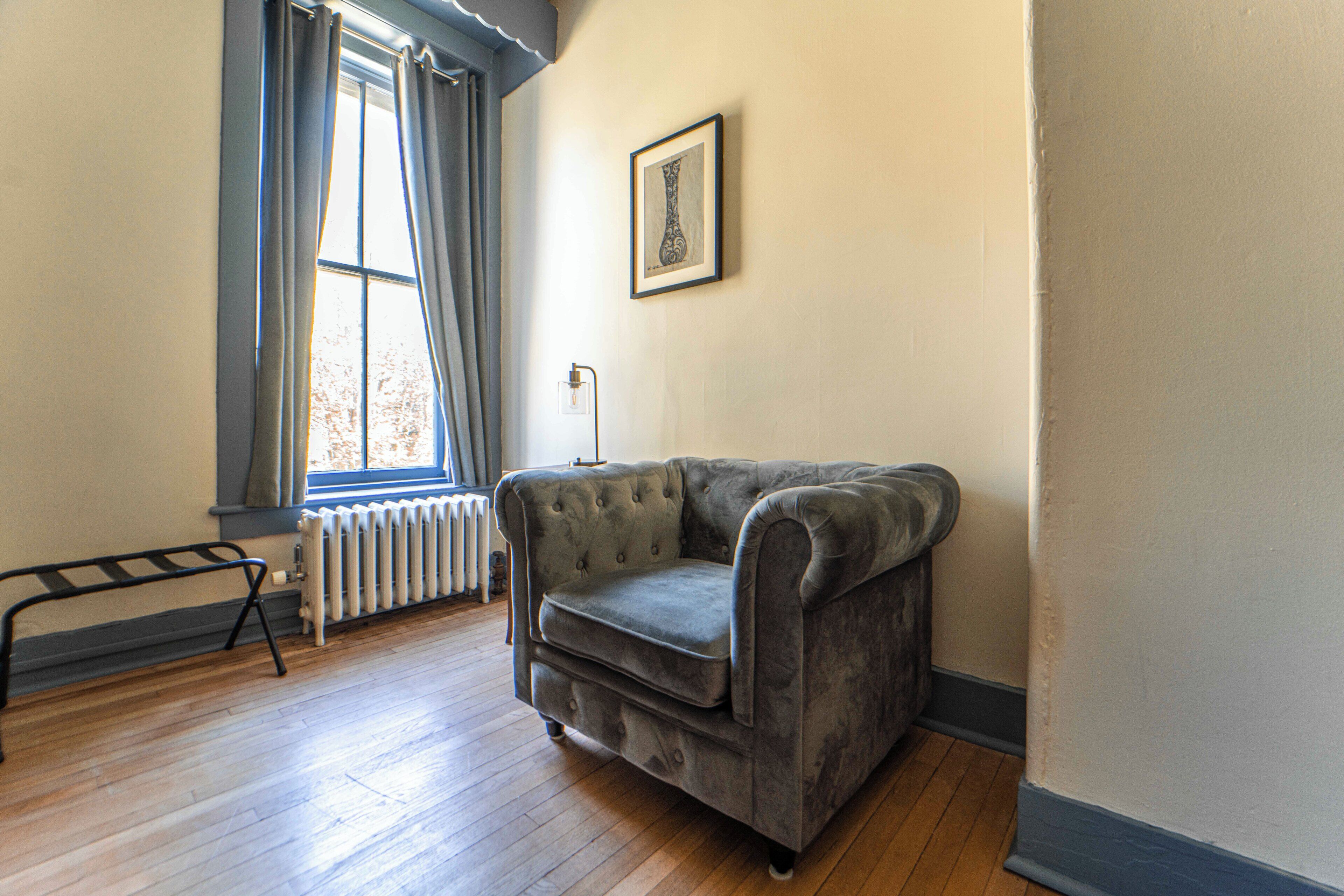 A room with a grey upholstered sofa, a radiator, a window with curtains, and a framed picture on the wall.