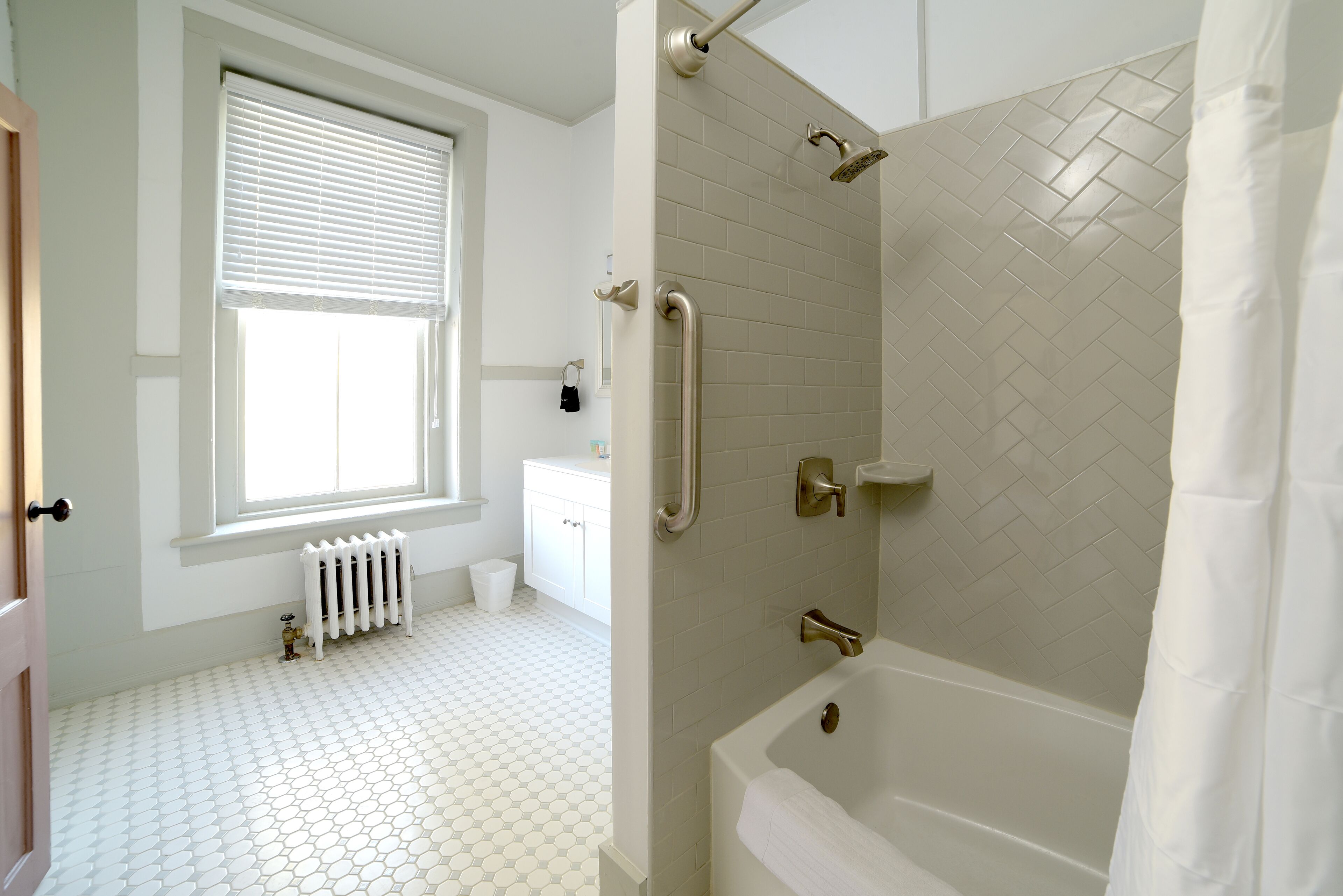 A bathroom with a white bathtub, a shower with a handheld showerhead, and a window with blinds.