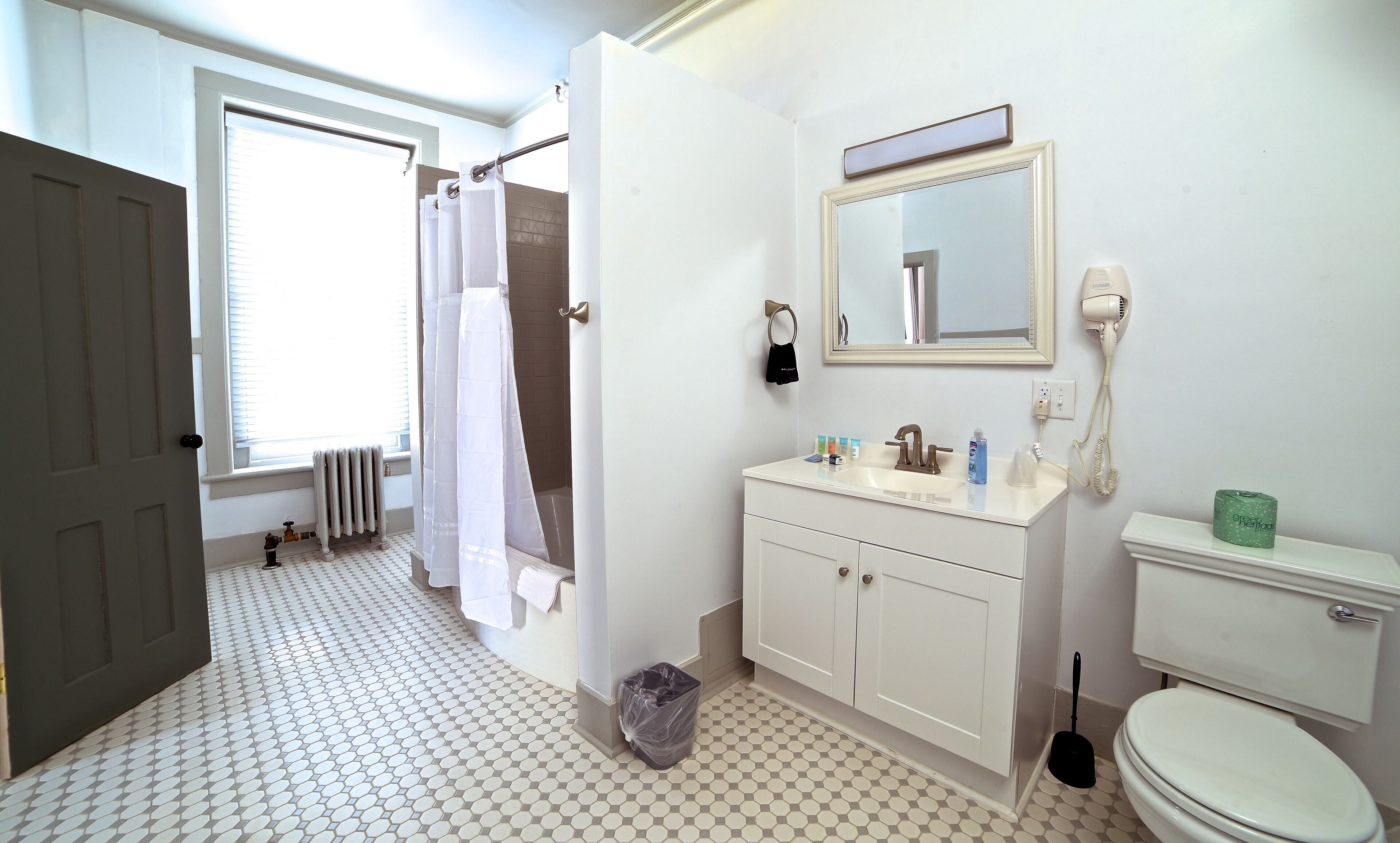 Un baño con inodoro, lavabo y espejo. Hay una cortina de ducha y un radiador cerca de la ventana.