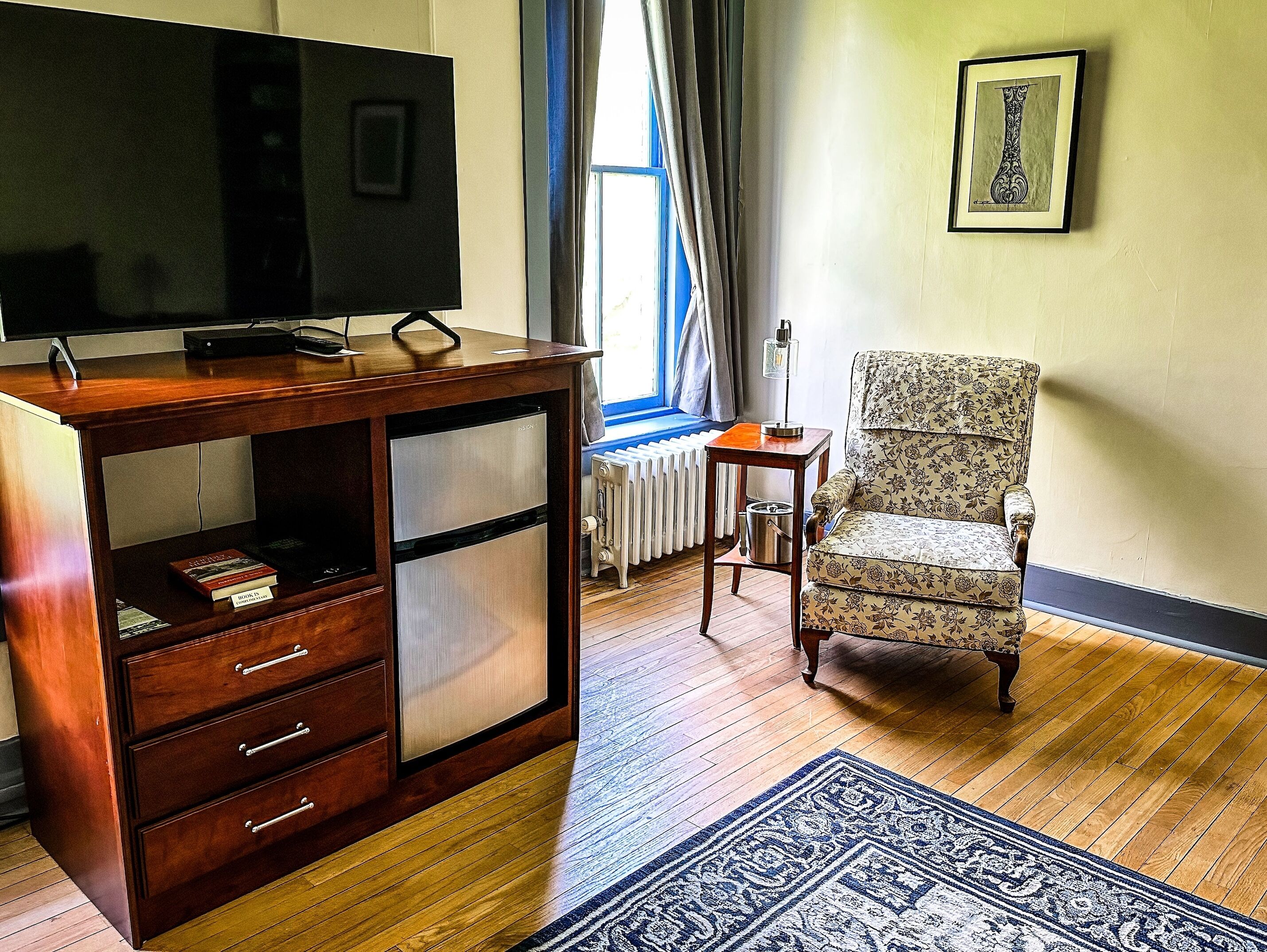A room with a wooden cabinet, a television on top, a patterned armchair, a small table with a lamp, and a framed artwork on the wall.