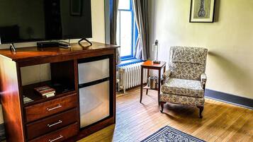 A room with a wooden cabinet, a television on top, a patterned armchair, a small table with a lamp, and a framed artwork on the wall.