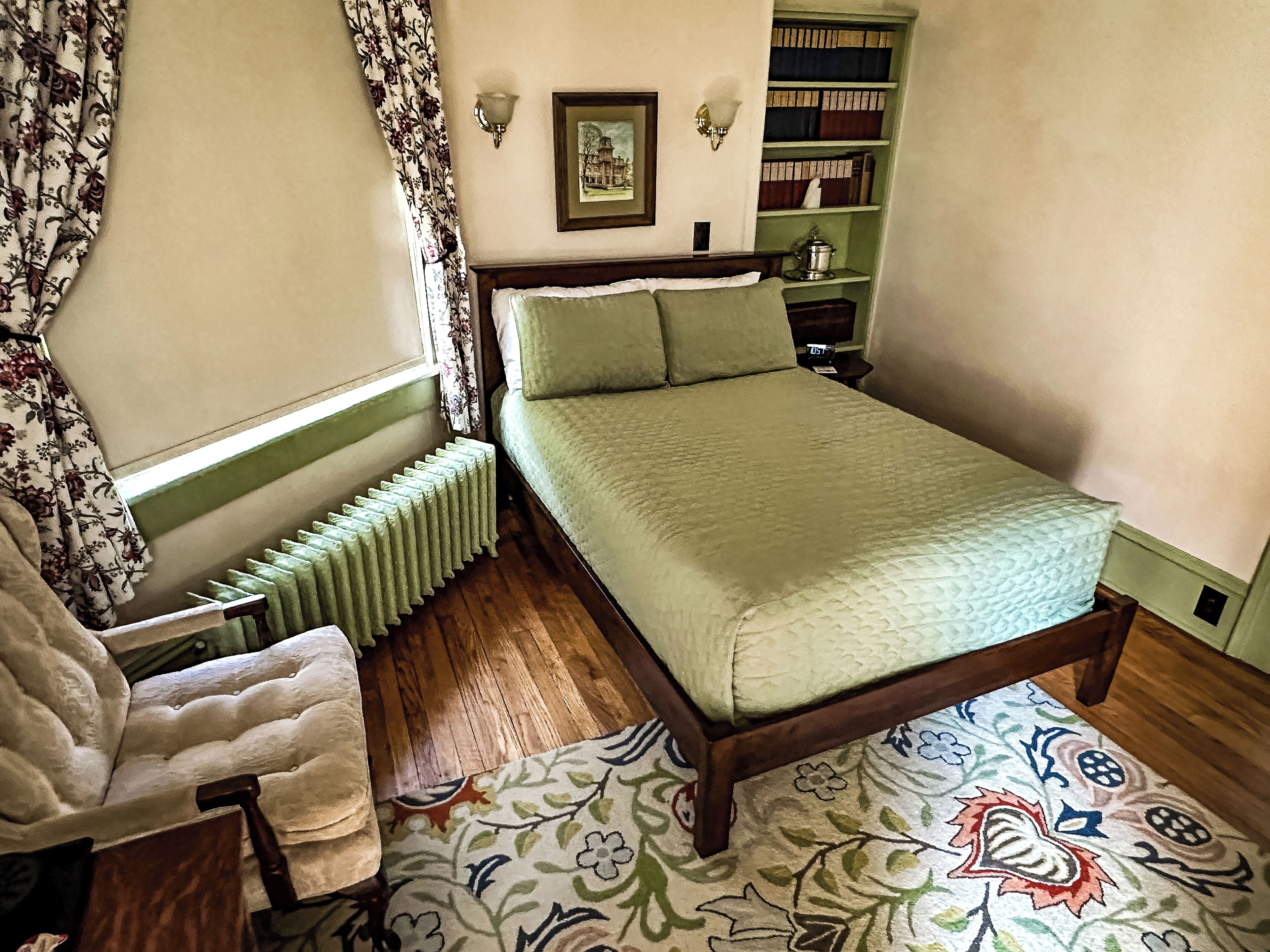 A bedroom with a bed, a chair, a radiator, a bookshelf, and a patterned rug.