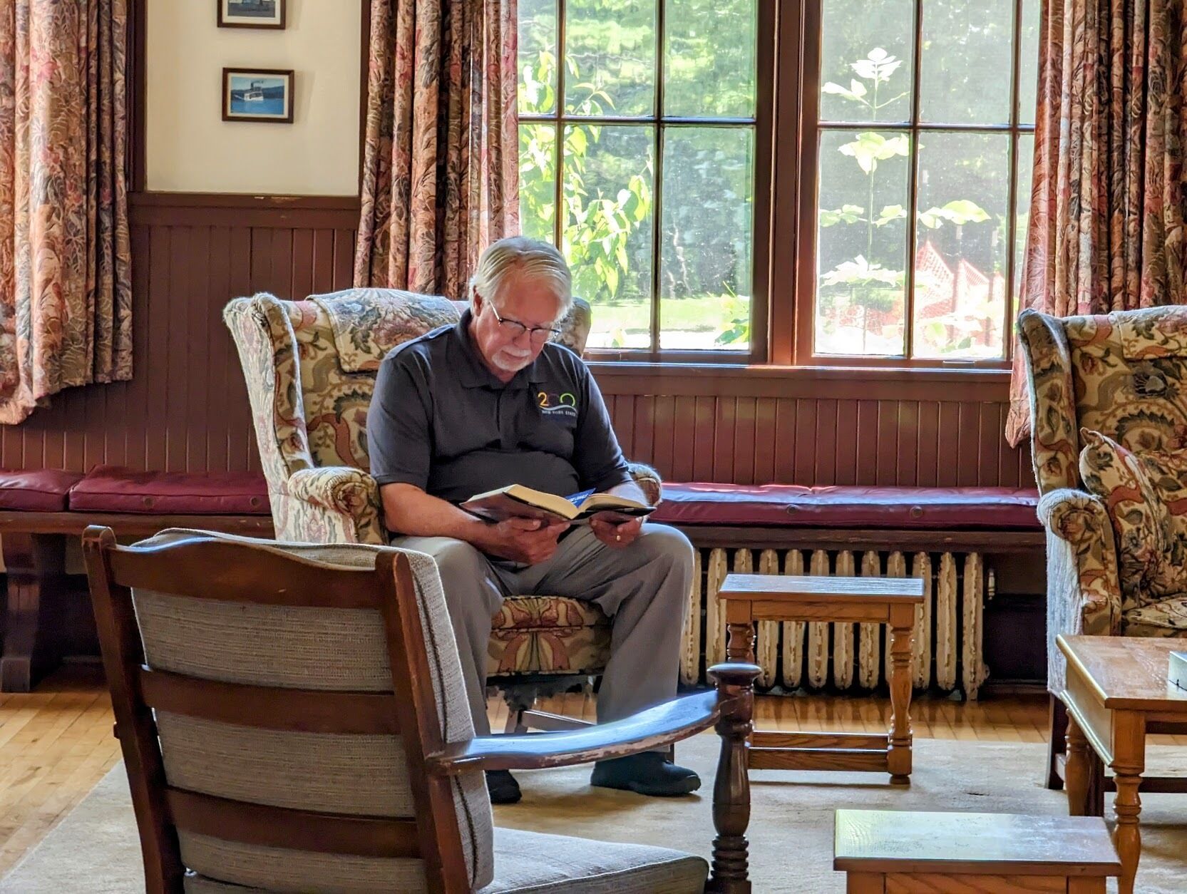 A man reading a book in a room with a wooden bench, patterned curtains, and a view of greenery outside.