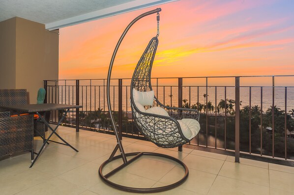 Un balcon agrémenté d’un fauteuil suspendu, d’une table et offrant une vue sur le coucher de soleil sur l’océan.
