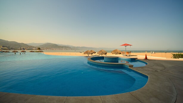 A large swimming pool with a curved design, surrounded by a concrete deck and a beach view. - WINTER OFFER!! CHIC & MODERN BEACHFRONT CONDO W/TERRACE (Puerto Vallarta)