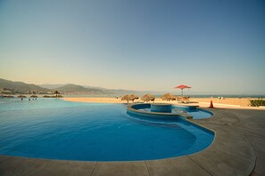 Une grande piscine à la forme incurvée, entourée d’une terrasse en béton et offrant une vue sur la plage.