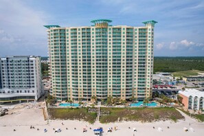 This beachfront resort features a striking high-rise building with distinctive green-tinted glass architectural elements, surrounded by lush coastal landscape and neighboring properties, with direct access to the sandy beach and ocean beyond. - Aqua 1710 - Beachfront Condo, Sleeps 6! (Panama City Beach)