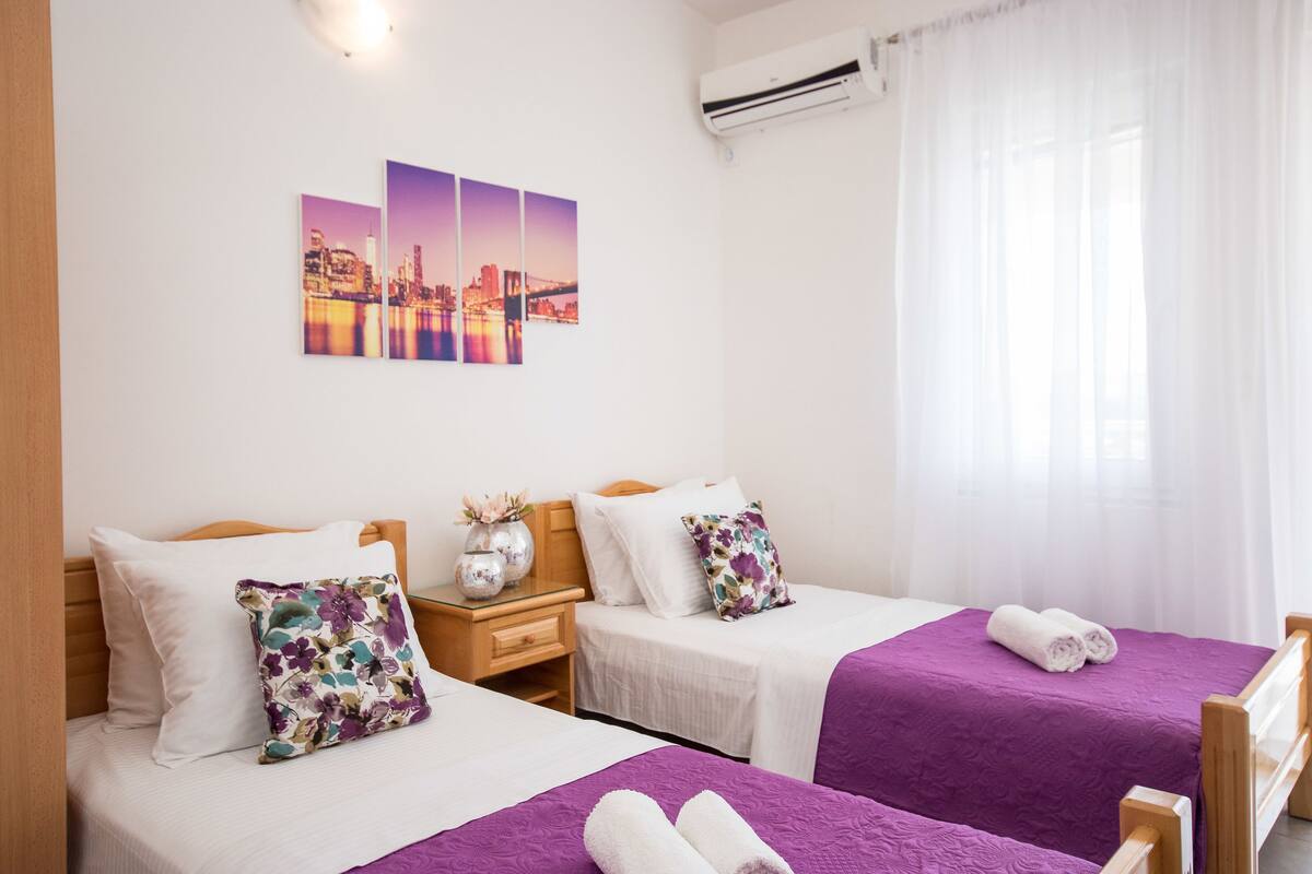 Habitación de hotel con dos camas, cada una con una colcha morada y almohadas blancas. Entre las camas, un mesita de noche con un florero y una lámpara. En la pared, sobre las camas, hay una obra de arte con un motivo urbano.