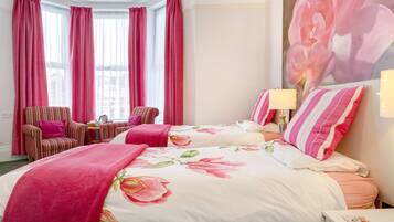 Une chambre d’hôtel avec deux lits, un fauteuil à rayures, une petite table avec une lampe et de grandes fenêtres ornées de rideaux roses.