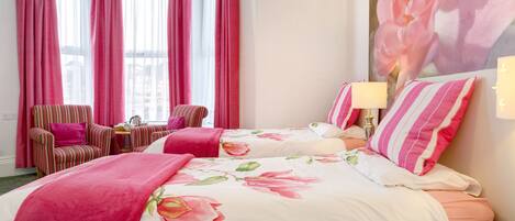 Ein Hotelzimmer mit zwei Betten, einem gestreiften Sessel, einem kleinen Tisch mit Lampe und großen Fenstern mit rosafarbenen Vorhängen.