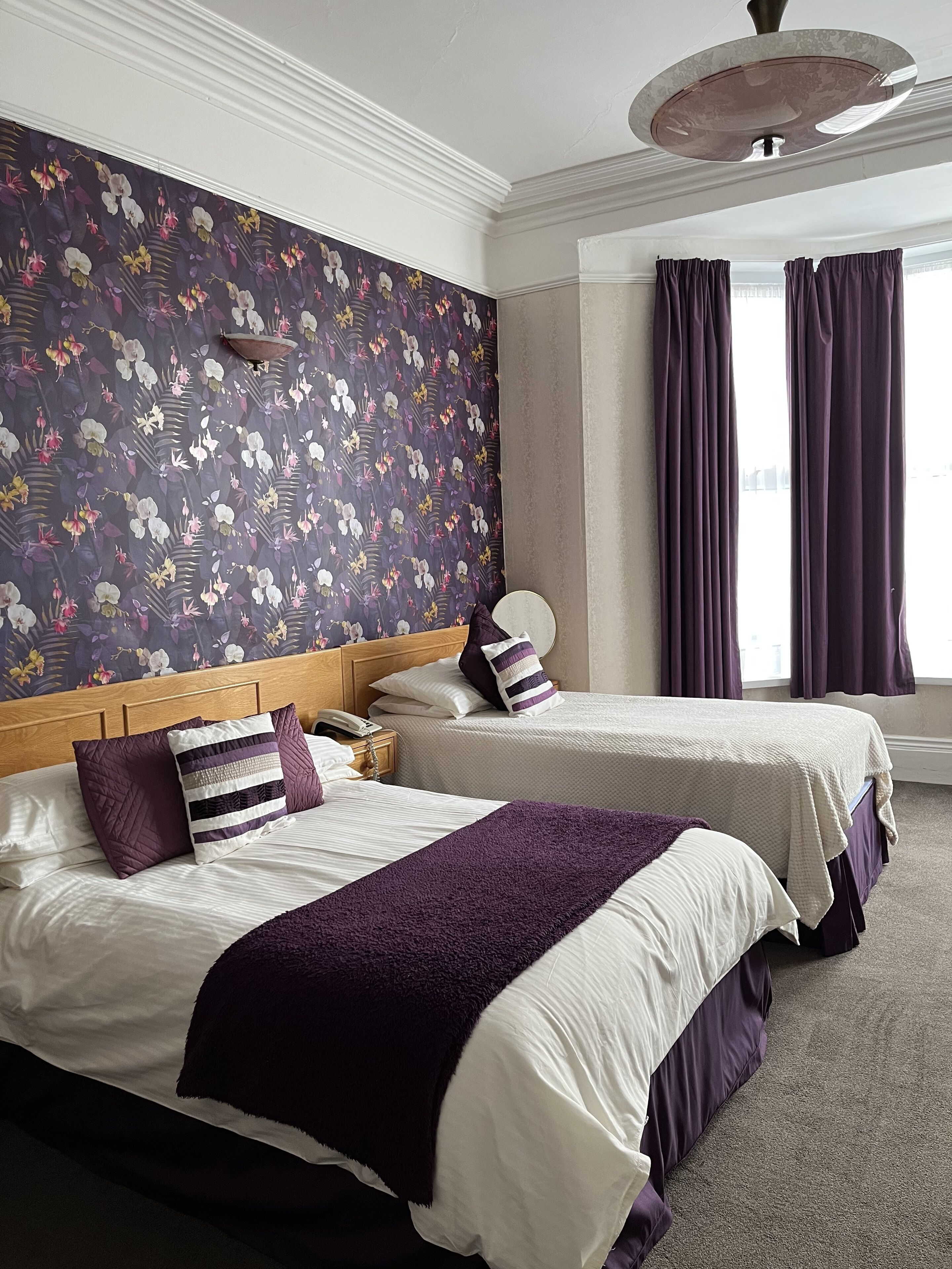 A hotel room with two beds, floral wallpaper, a wooden headboard, a round light fixture, and a window with curtains.
