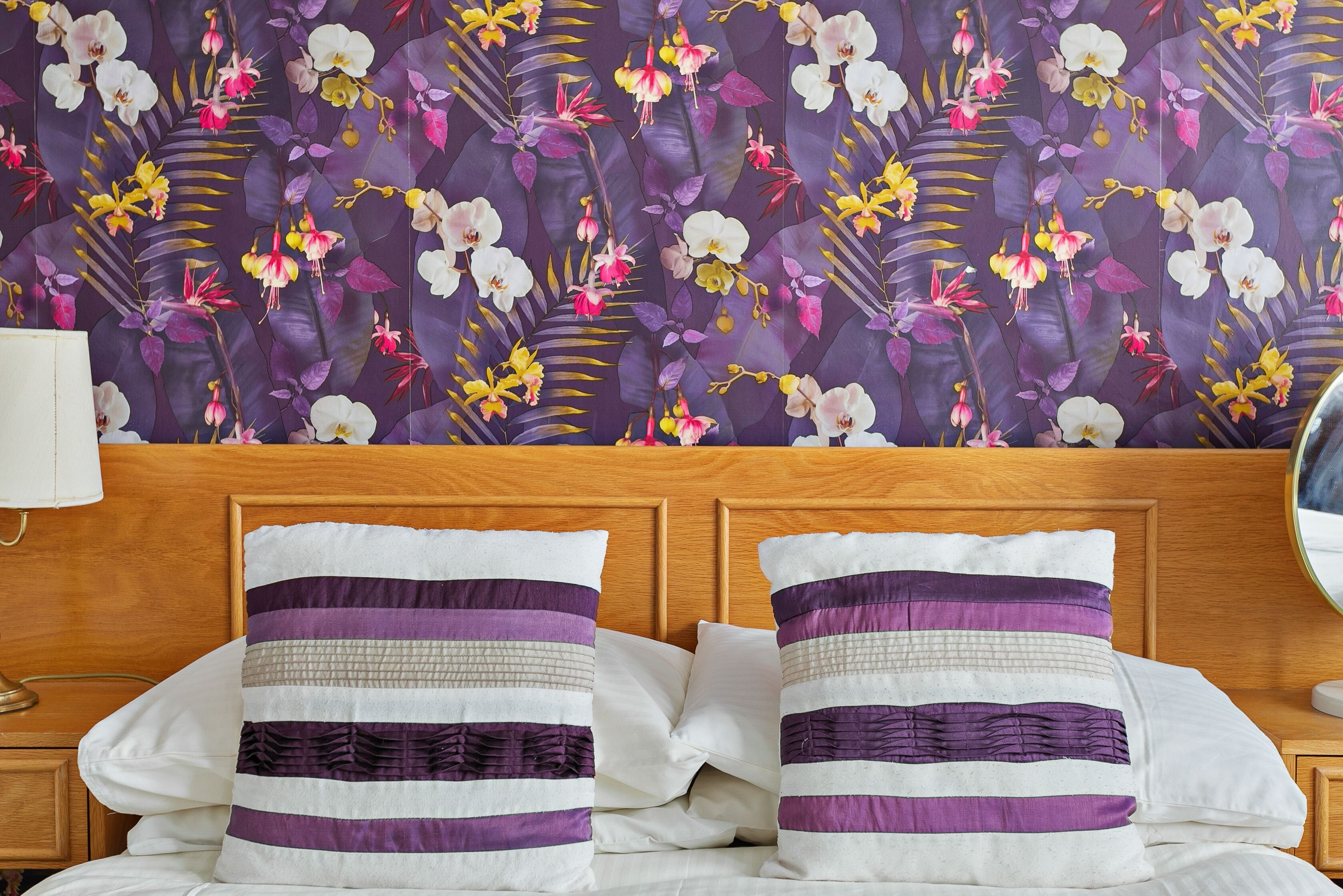 Habitación de hotel con cabecera de madera, dos almohadas estampadas con rayas y papel tapiz floral.