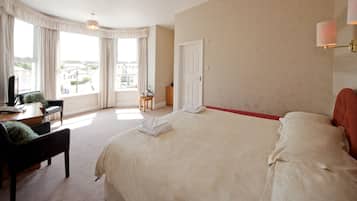 Habitación de hotel con una cama grande, un escritorio con una silla, una mesita y vistas al exterior a través de amplios ventanales.