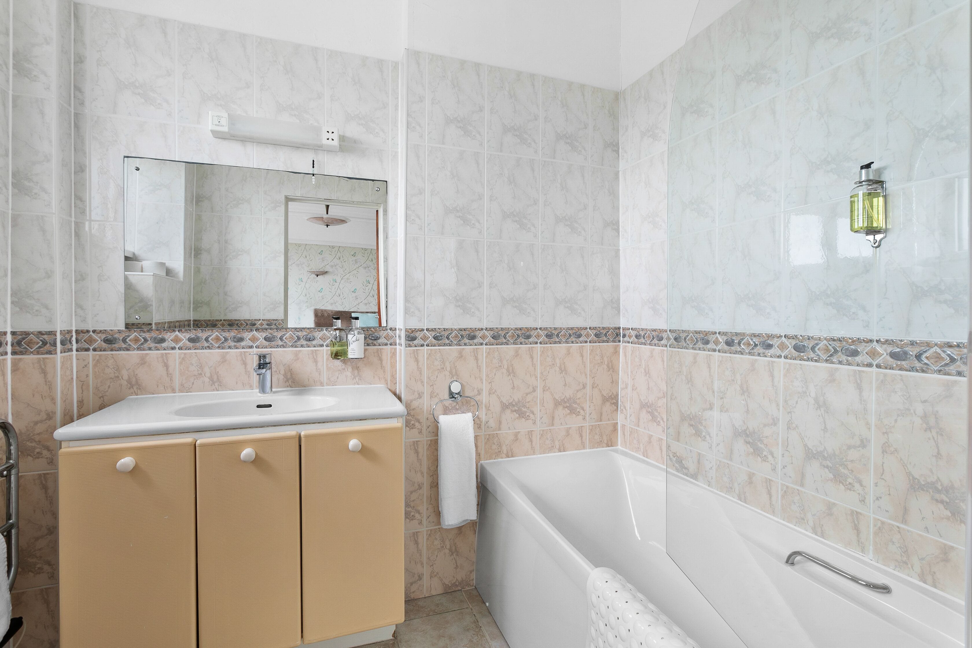 A bathroom with a white sink, beige cabinets, a white bathtub, and a mirror with a wall-mounted light.