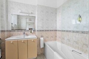 A bathroom with a white sink, beige cabinets, a white bathtub, and a mirror with a wall-mounted light. - Marine Hotel Paignton (Paignton)