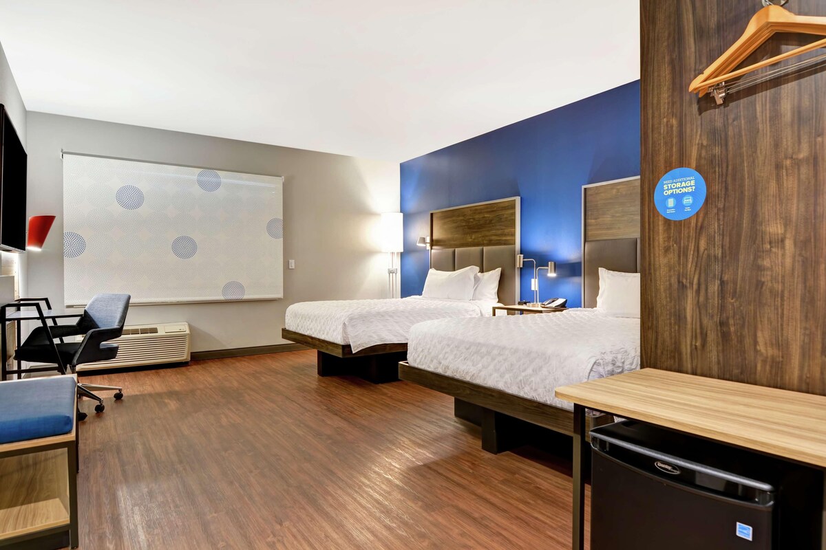 A hotel room with two beds, a desk, a chair, a heater, and a wall-mounted TV.