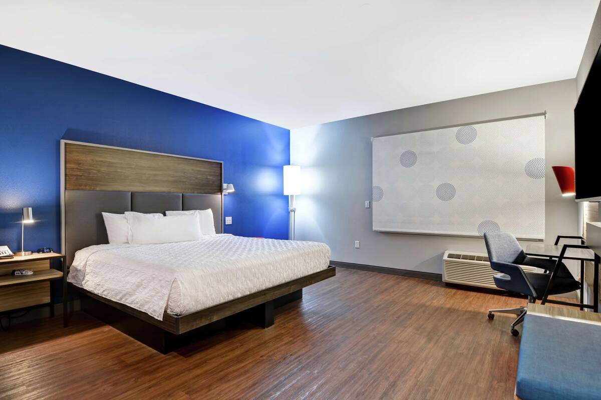 A hotel room with a bed, a desk, a chair, a lamp, and a wall with a geometric pattern.