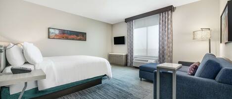 Quarto de hotel com cama, sofá, televisão, escrivaninha e janela com cortinas.