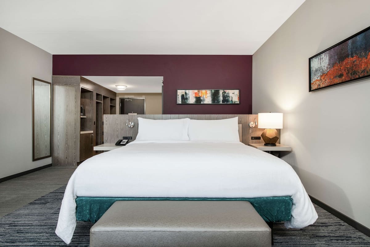 Ein Hotelzimmer mit einem großen Bett, einem Schreibtisch, einem Stuhl, einem Nachttischchen mit Lampe, einem Bild an der Wand und einem Kleiderschrank.