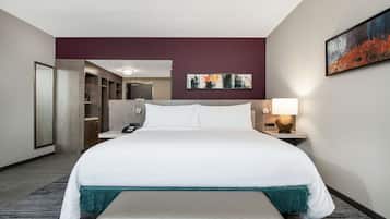 A hotel room with a large bed, a desk, a chair, a bedside table with a lamp, a painting on the wall, and a wardrobe.