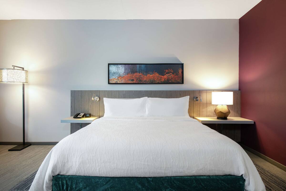 Ein Hotelzimmer mit einem großen Bett, einem Nachttisch mit Lampe und einem Gemälde an der Wand.