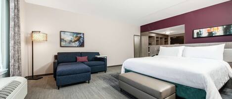 A hotel room with a large bed, a sofa, a bench, a standing lamp, and artwork on the walls.