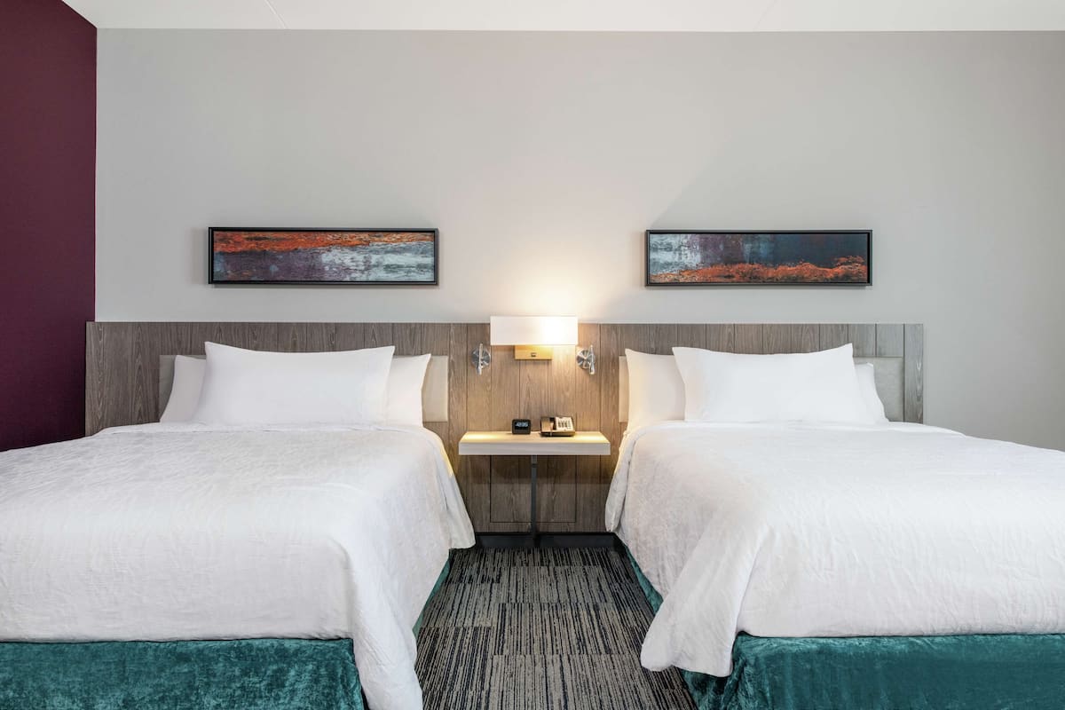 Ein Hotelzimmer mit zwei Betten, einem Nachttisch, einer Lampe und zwei gerahmten Kunstwerken an der Wand.