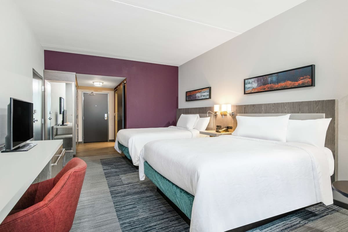 Ein Hotelzimmer mit zwei Betten, einem Schreibtisch mit Fernseher, einem roten Sessel und einer Tür zu einem Badezimmer.