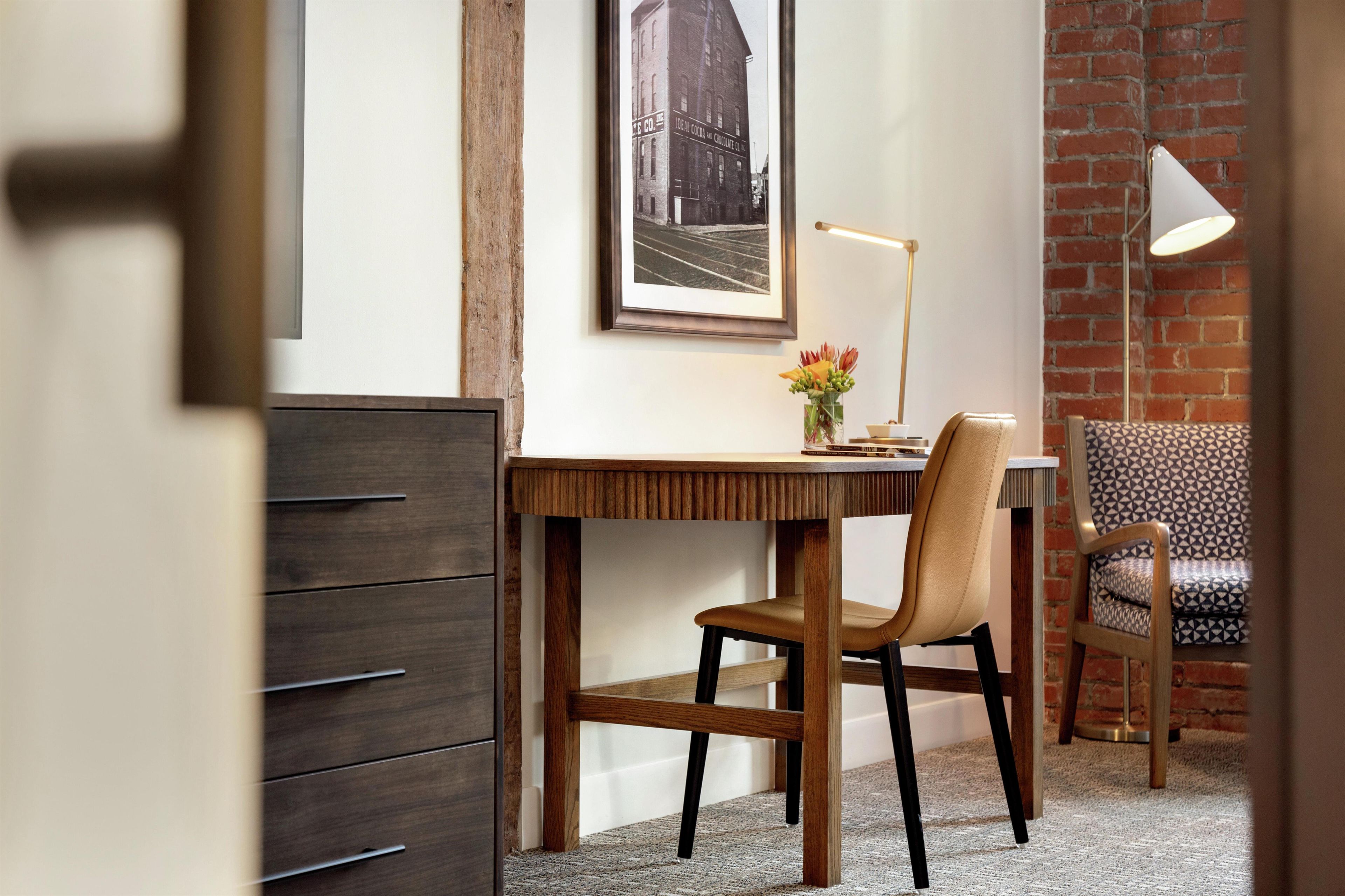 A room with a wooden desk, a chair, a framed picture on the wall, and a patterned chair in the background.